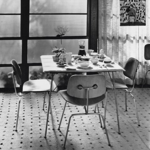 Eames DCM Molded Wood Dining Chair with Metal Base around a table, archival imagery.