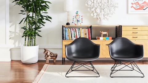 Duas poltronas Eames em plástico moldado, com base aramada, sobre um tapete em um ambiente de sala de estar.