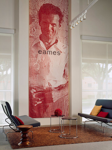 Dois cinzentos Eames Sofa Compacts frente a frente em um lobby de escritório com uma grande parede de um jovem Charles Eames.