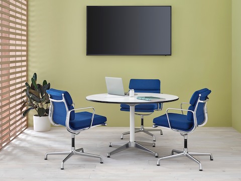 Uma pequena área de reunião com uma mesa redonda Eames com tampo branco e três Eames Soft Pad Cadeiras com estofamento azul.