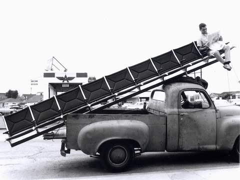 Vintage photo of a man sitting at the end of an Eames Tandem Sling Seating run being delivered via pickup truck. 