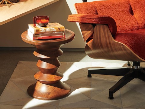 Uma banqueta Eames Turned C com acabamento em nogueira junto a uma Lounge Chair Eames com tecido laranja queimado.