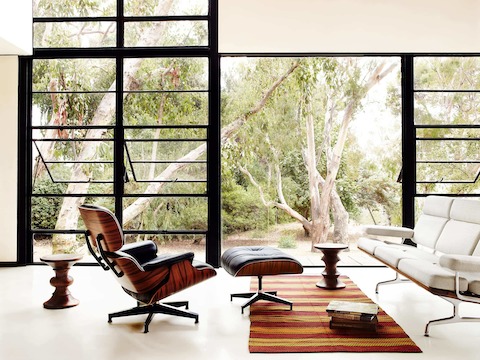 Two walnut Eames Turned Stools, an Eames Lounge Chair and Ottoman, and a white Eames Sofa in an open environment with large windows. 