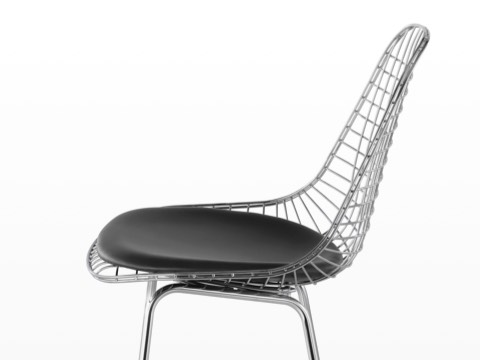 La mitad superior de un taburete de alambre Eames plateado con una almohadilla de asiento negra, vista desde un lado.