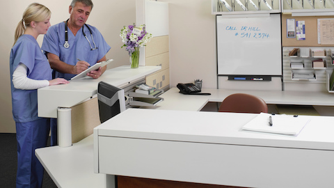 Two healthcare workers review a chart while standing next to a transaction surface on an Ethospace Nurses Station. 