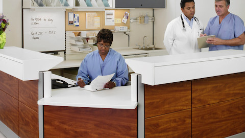 Tres profesionales de la salud consultan cuadros en una estación de enfermeras Ethospace con azulejos de madera.