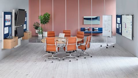 Seis Eames Soft Pad Chairs de color naranja quemado rodean una mesa en forma de lágrima en un espacio de colaboración con elementos de visualización Exclave.