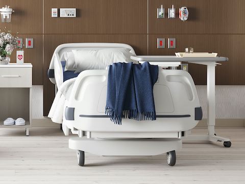 A patient room with a hospital bed in front of a headwall in a walnut finish with a Nemschoff Bedside Cabinet next to it and an EZ-123 Overbed Table on the other side.