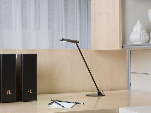 A black freestanding Flute Personal Light provides targeted illumination on a desktop. A black freestanding Flute Personal Light provides targeted illumination on a desktop.