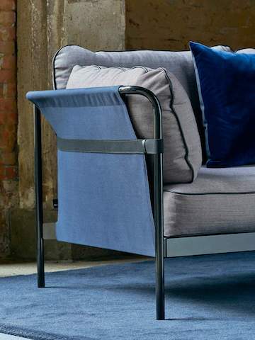 The side of a Can Sofa from HAY in light gray fabric upholstery and a black frame with blue sling, shown in a loft setting with exposed brick walls.
