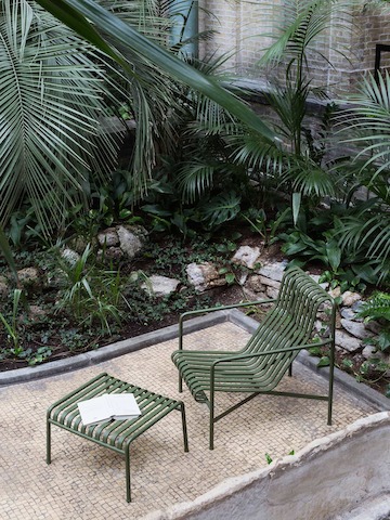 An olive green Palissade Lounge Chair and Ottoman on a small patio surrounded by ferns and a garden.