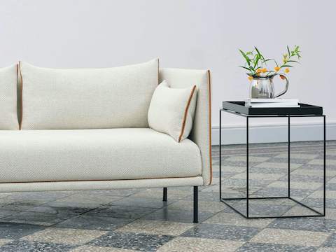 A Silhouette Sofa from HAY in off-white upholstery with tan piping and black legs sitting beside a square, black Tray Side Table.