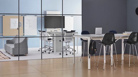 Cadeias em fibra de vidro moldadas Eames, pretas, em uma mesa Layout Studio com altura para trabalhar em pé, com vista de outra sala com mesa com altura para trabalhar em pé e banquetas Setu.