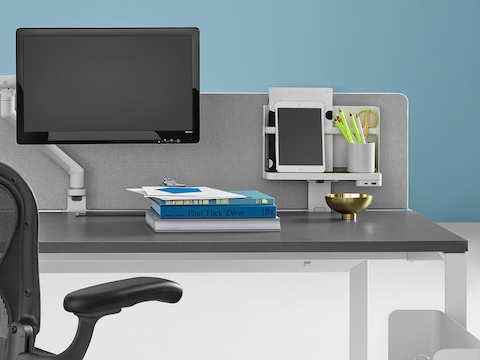 Close-up of a Layout Studio bench with a gray flat-edge fabric, center privacy screen, graphite Aeron Chair, Flo Monitor Arm, and Ubi Mobile Bag Catch.