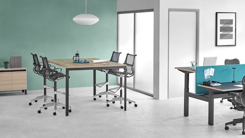 Agencement de bureau avec une table haute Layout Studio et des sièges hauts Setu noirs, adjacents à un bureau Ratio avec un écran de séparation bleu et des sièges Mirra 2 noirs.