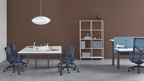 Büroumgebung mit einer Layout Studio-Bench für vier Personen und dunkelblauen Cosm-Stühlen neben einem höhenverstellbaren Ratio-Schreibtisch mit einer blauen Trennwand und einem dunkelblauen Cosm-Stuhl.