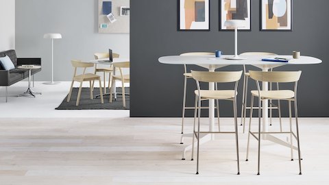 Quatro banquetas Leeway com bases de metal e assentos e encosto em madeira clara, em uma mesa para trabalhar em pé, perto de um lounge com um sofá e cadeiras pretas junto a uma mesa.