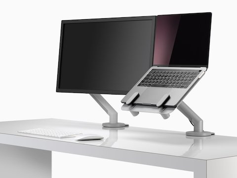 Tela de monitor e um laptop aberto e elevado na linha dos olhos, apoiado por um suporte para laptop Ollin e um braço para monitor Flo.
