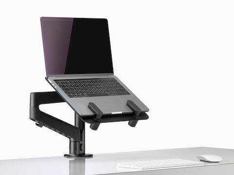 Vista en ángulo lateral de una portátil abierta, elevada sobre un escritorio, sujetada mediante un soporte para computadora portátil Lima en negro y un brazo articulado para monitor Lima.