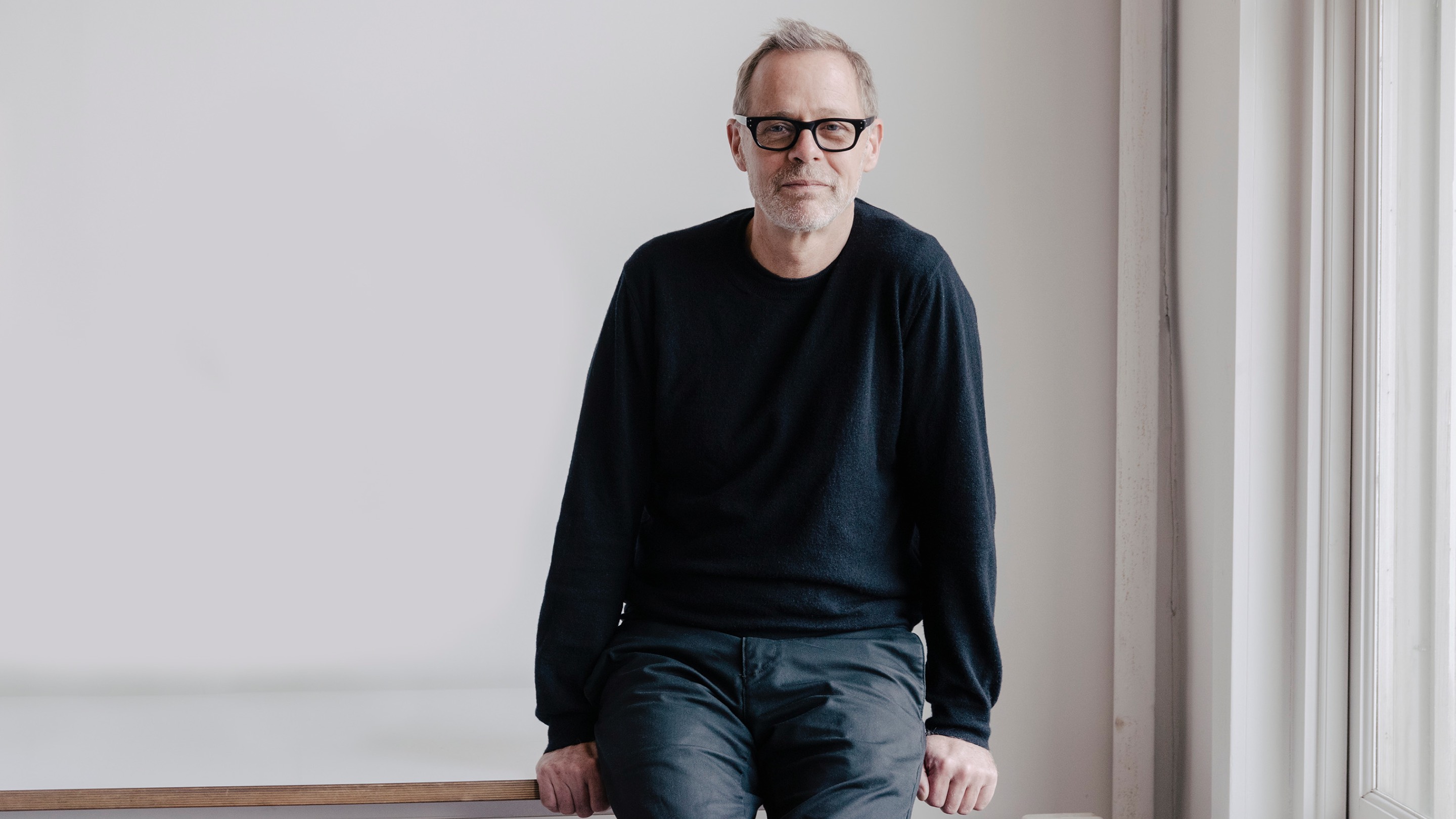 Portrait of designer Neil Logan perched on a white table and white background.