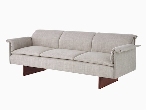 Visão angular de um sofá de três lugares Mantle estofado em Capri Stone com base em madeira de nogueira e almofadas sobrepostas.