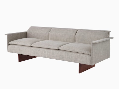 Vista en ángulo de un sofá de tres asientos Mantle tapizado en Capri Stone con base de madera de nogal.