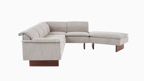 Vista lateral de un sofá de tres asientos Mantle combinado con un descansapies Mantle en tapicería Capri Stone con base de madera de nogal.