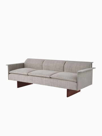 Vista en ángulo de un sofá de tres asientos Mantle tapizado en Capri Stone con base de madera de nogal.