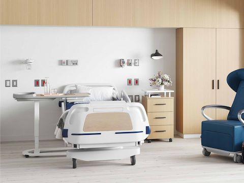 Mirage Overbed Table over a hospital bed in a patient room, Bedside Cabinet and Mora wardrobe in an ash finish next to it, and Ava Recliner in blue upholstery nearby.