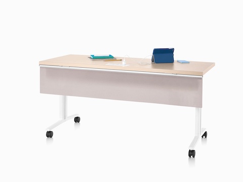 Viewed at an angle, an Everywhere Table with a clear on ash woodgrain top, fabric modesty panel attached to the surface and a white base with casters.