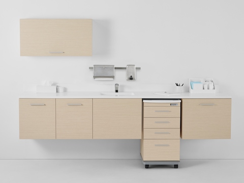 A Mora casework wall in a light ash finish consisting of one overhead storage cabinet and three lower supply cabinets with a supply cart docked within the casework.