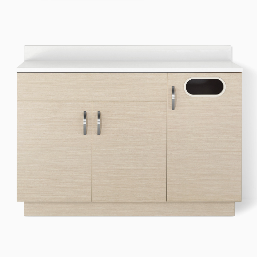 A Mora Casework lower storage cabinet in an ash wood finish with double doors, a trash cabinet, and a white solid surface countertop.