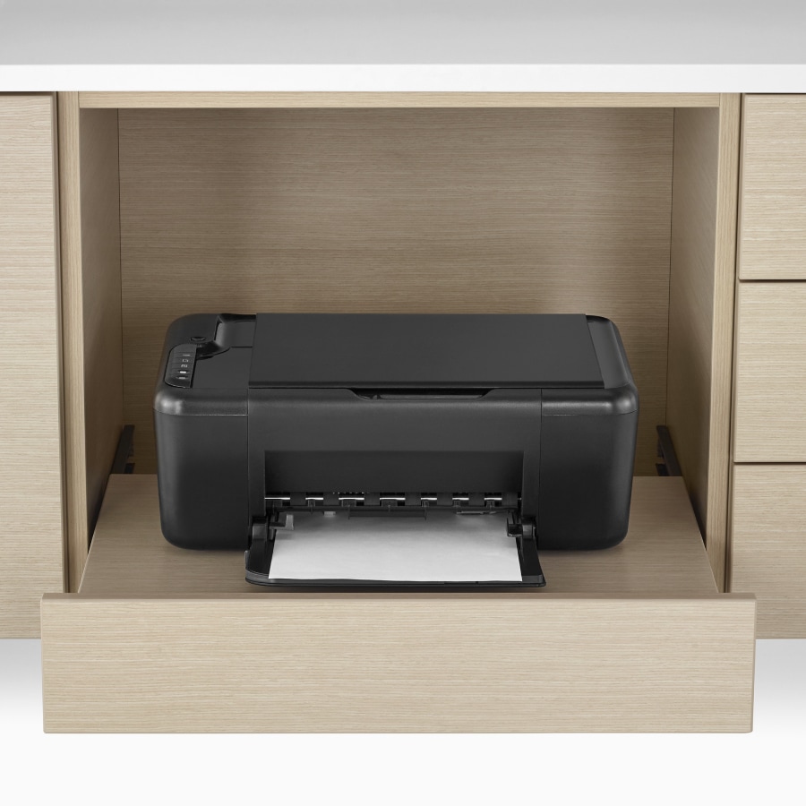 A close-up view of a Mora Casework technology storage cabinet in an ash finish holding a black printer.