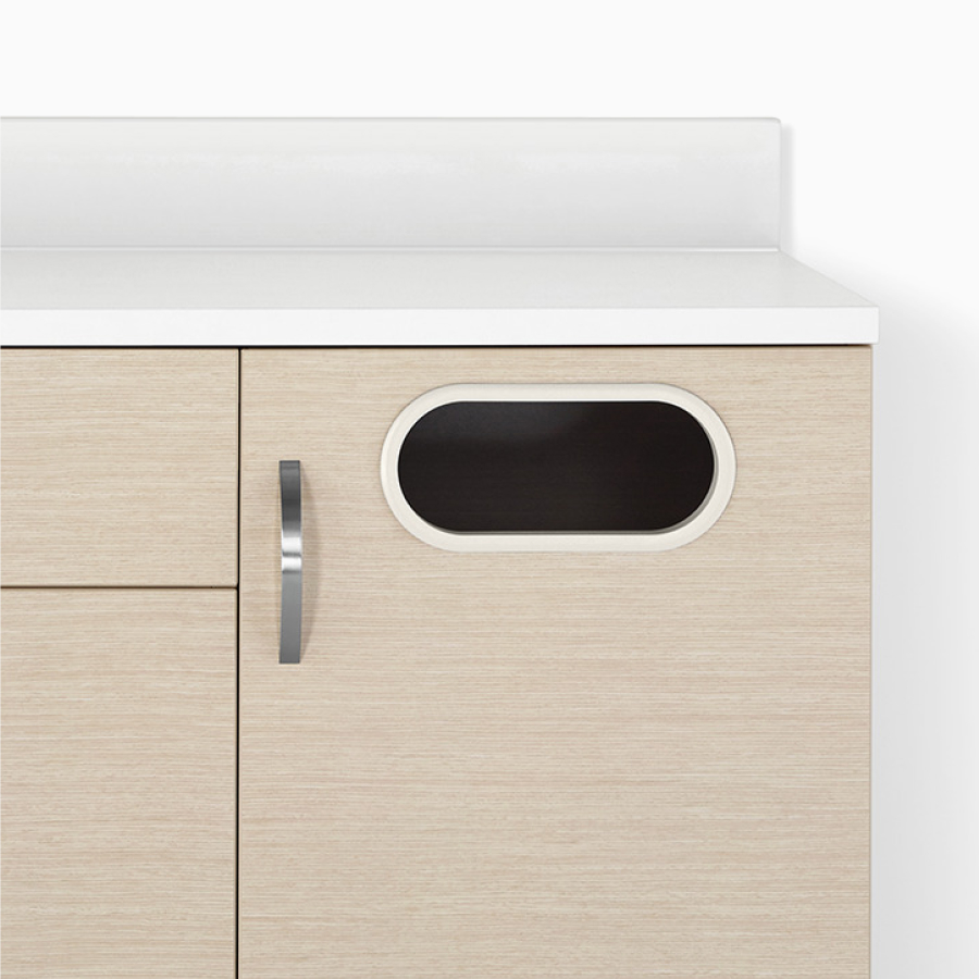 A close-up view of a Mora Casework trash receptacle cabinet in an ash wood finish with a white solid surface.