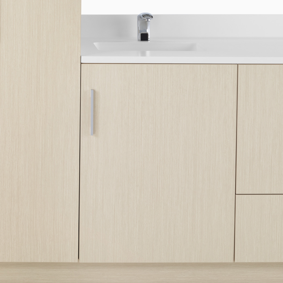 A close-up view of Mora Casework in an ash wood finish with a storage case door and a white solid surface top with integrated sink.