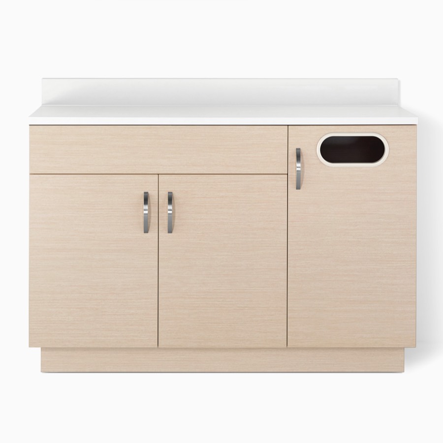 A Mora System casework lower storage cabinet in an ash wood finish with double doors, a trash cabinet, and a white solid surface countertop.