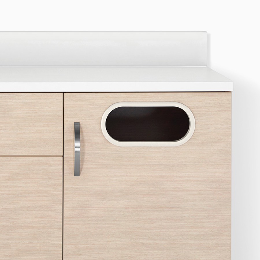 A close-up view of a Mora System casework trash receptacle cabinet in an ash wood finish with a white solid surface.