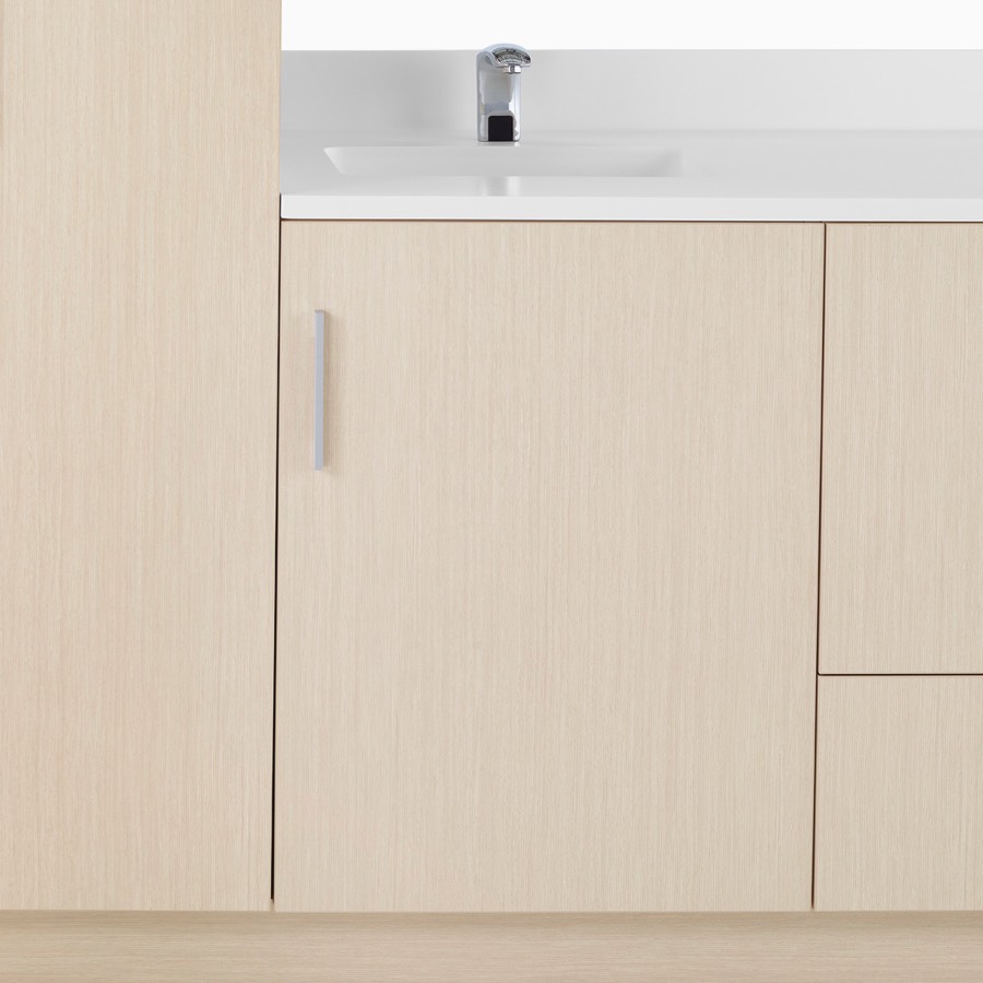 A close-up view of Mora System casework in an ash wood finish with a storage case door and a white solid surface top with integrated sink.