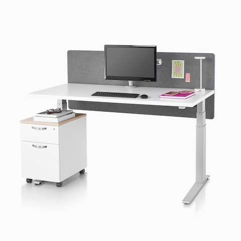 Standing desk, Motia Sit-to-Stand Table, with a white top, metallic silver base, surface-attached privacy screen, desk lamp, and a Tu Wood pedestal. Standing desk, Motia Sit-to-Stand Table, with a white top, metallic silver base, surface-attached privacy screen, desk lamp, and a Tu Wood pedestal.