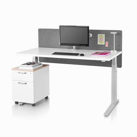 Standing desk, Motia Sit-to-Stand Table, with a white top, metallic silver base, surface-attached privacy screen, desk lamp, and a Tu Wood pedestal.