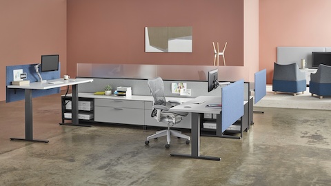 Una estación de trabajo de altura ajustable Motia con un escritorio en blanco, pantalla de privacidad sujetable a superficies en gris, y un monitor digital montado en un brazo articulado.
