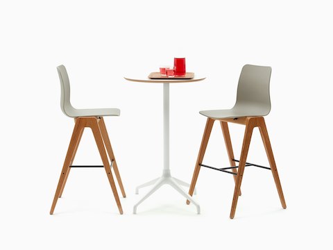 Una mesa Ali con altura de bar, con base de estrella de 4 puntas en blanco y superficie de chapa de madera circular en roble, con dos taburetes con altura de bar Polly.