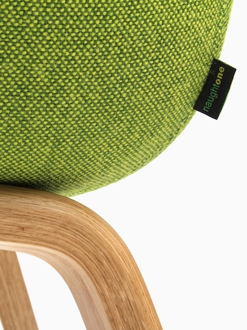 Detalle en primer plano de una etiqueta NaughtOne en la esquina posterior izquierda de una silla de visita Always, tapizada en tela verde texturada y montada sobre una base de madera.
