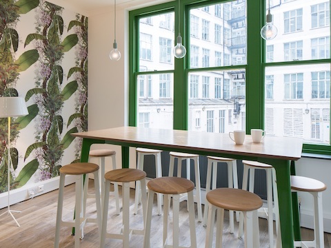 Table de bar Fold de NaughtOne avec plateau à placage bois vernis et piètement vert devant une fenêtre entourée de tabourets Construct blancs.