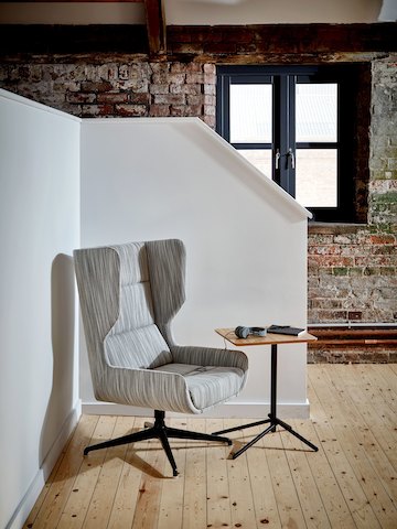 Un sillón Hush de NaughtOne en estampado gris claro, ubicado en una esquina con una mesa lateral Knot de superficie de roble y base negra.