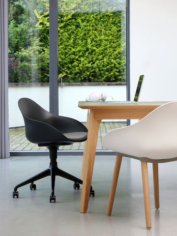 Una silla Ruby en negro combinada con una base de estrella de 5 puntas y asiento acolchado en gris y ruedas giratorias que se encuentra junto a una silla de visita Ruby en blanco sobre una base de clavijas en roble y una mesa.