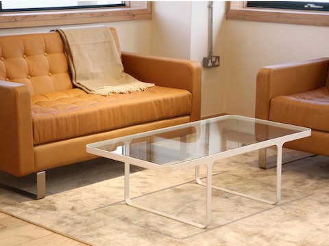 Una mesa de café Trace de NaughtOne blanca con superficie de vidrio, frente a un sofá y una silla club Clyde de cuero.