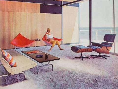 A woman sits in one of a pair of orange-pink Nelson Coconut Chairs in the sunny living room of a mid-century modern home in Malibu. An Eames Lounge Chair and Ottoman and Eames Sofa Compact are nearby. 
