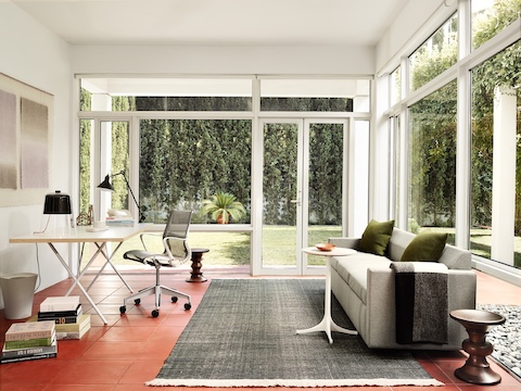 Una mesa de pedestal Nelson blanca sirve como una pieza de acento funcional en una oficina en casa con paredes de vidrio con una vista al aire libre.