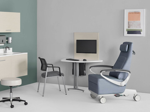 An exam room setting with Mora System casework on the wall in a light wood finish and a Mora peninsula on the side wall located between a Nemschoff Ava patient recliner and a Verus side chair.